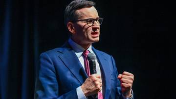 Mężczyzna w niebieskim garniturze i okularach przemawia do mikrofonu.