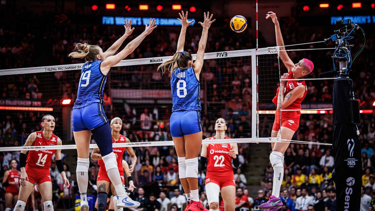 Siatkarka w czerwonej koszulce blokuje piłkę lecącą do siatki, podczas gdy dwie siatkarki w niebieskich koszulkach skaczą do jej odbicia. Widzowie na trybunach.