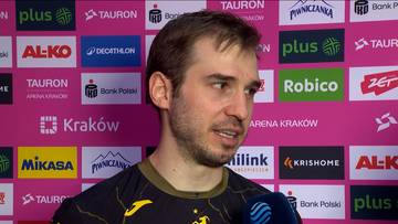 Mężczyzna z zarostem i kr&oacute;tkimi włosami w ciemnej koszulce, na tle ściany z logo sponsor&oacute;w.