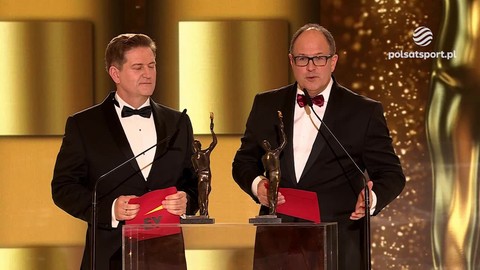 Dwóch mężczyzn w smokingach z muszkami, każdy z czerwoną kartką i statuetką, stoi za mównicami podczas gali wręczenia nagród.