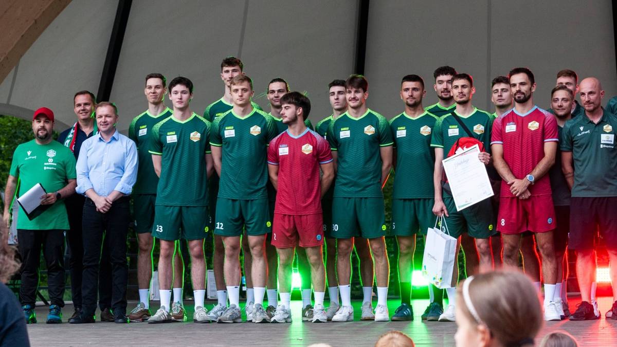 Grupa zawodnik&oacute;w w strojach sportowych, część w zielonych, część w czerwonych, stoi na scenie.