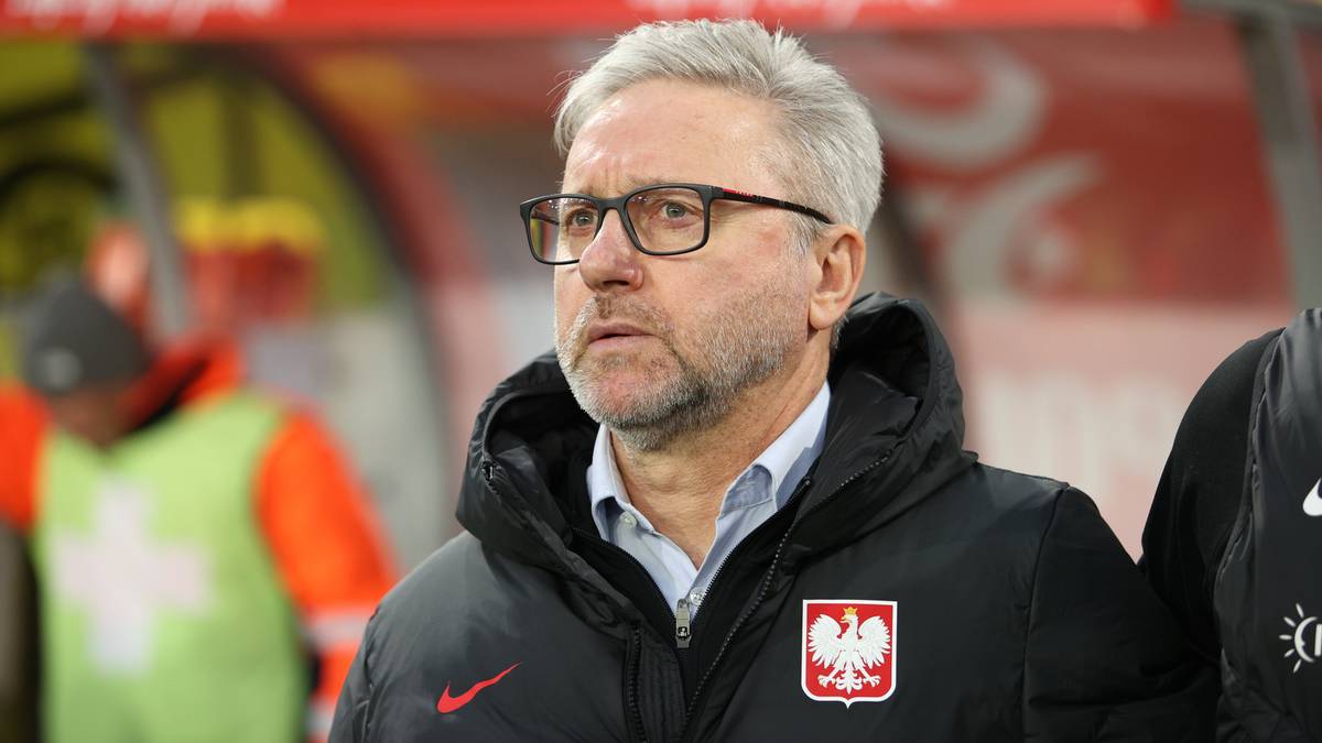 Portret mężczyzny z siwymi włosami i brodą, noszącego okulary i kurtkę z herbem Polski.