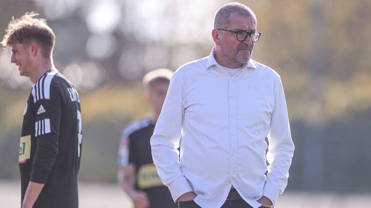 Starszy mężczyzna w okularach i białej koszuli stoi z rękami w kieszeniach, z innymi osobami z drużyny piłkarskiej w tle.