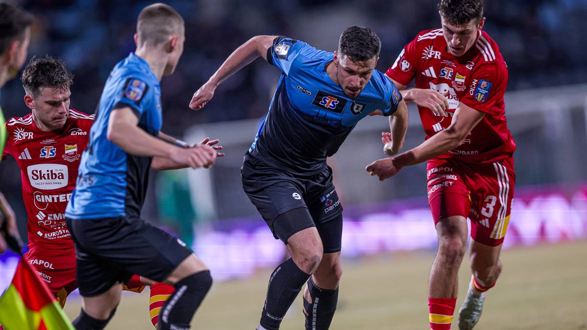 Trzech piłkarzy podczas meczu, jeden w niebiesko-czarnej koszulce prowadzi piłkę, dw&oacute;ch w czerwonych strojach pr&oacute;buje go zatrzymać.