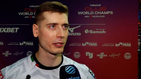 Portret mężczyzny w białej koszulce z kolorowymi elementami, trzymającego mikrofon. W tle widnieje ściana z logo "VOLLEYBALL CLUB WORLD CHAMPS" i sponsorami.