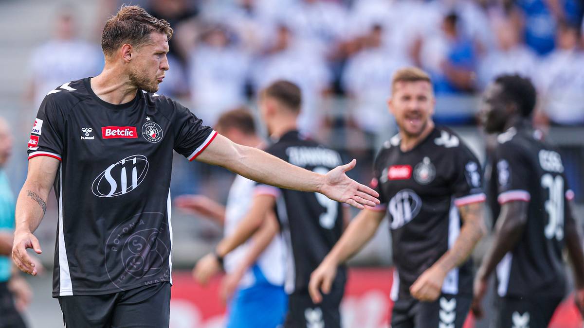 Piłkarz w czarnej koszulce z białymi elementami i logo "Betclic" gestykuluje ręką, patrząc w bok. W tle inni zawodnicy i widzowie.