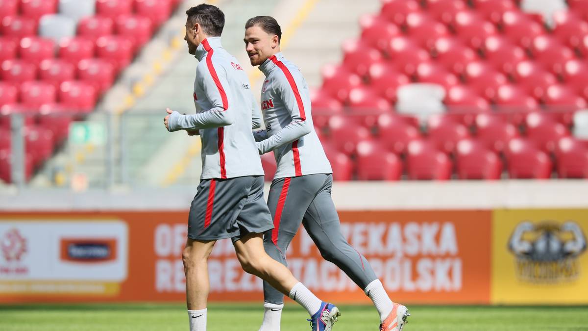 Dw&oacute;ch piłkarzy w szarych strojach reprezentacji Polski biegnie po zielonej murawie stadionu.