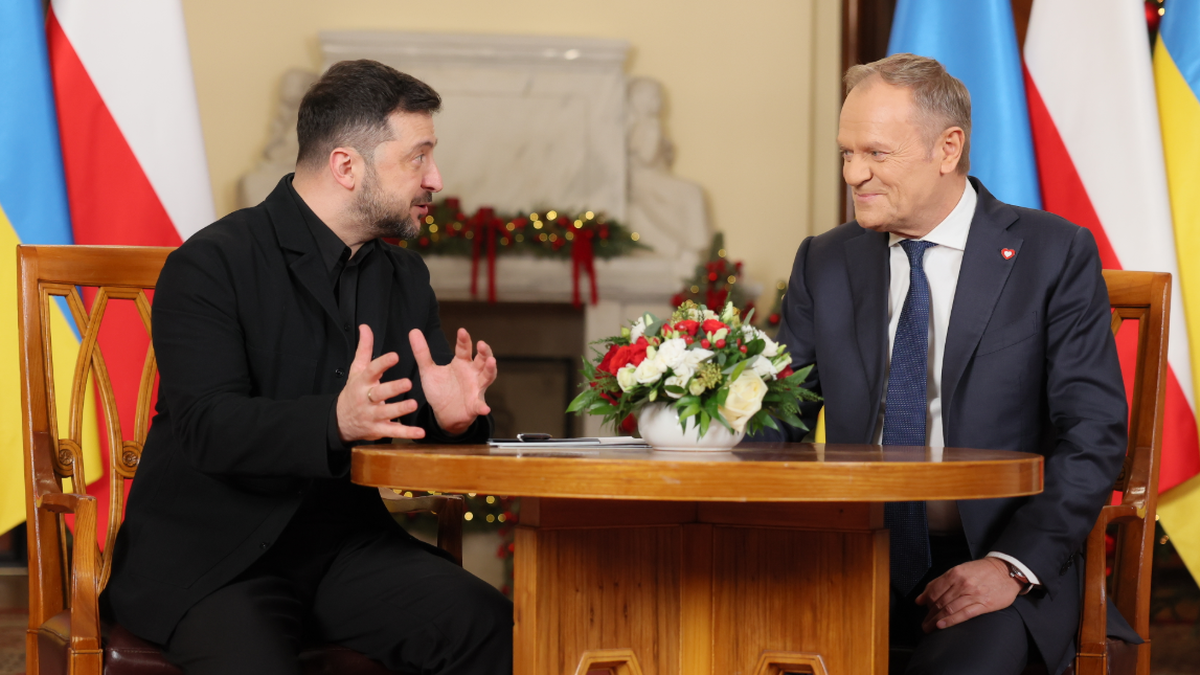 Premier Donald Tusk rozmawia z prezydentem Ukrainy Wołodymyrem Zełenskim przy stole, w tle flagi Polski i Ukrainy.