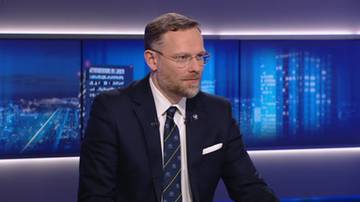 Zbigniew Bogucki w garniturze i krawacie siedzi przed ekranami wyświetlającymi panoramę miasta.