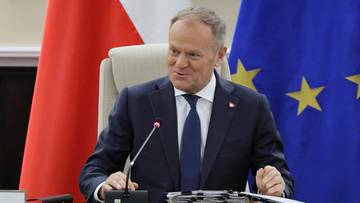 Premier Donald Tusk siedzi przy stole, za nim polska i europejska flaga.