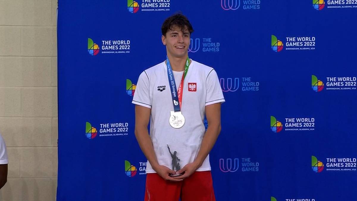 Młody mężczyzna w białej koszulce z logo The World Games i medalem na szyi, trzymający puchar.