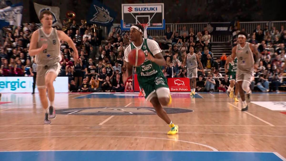 Koszykarz w zielonym stroju z piłką biegnie po boisku, w tle inni zawodnicy i kibice.