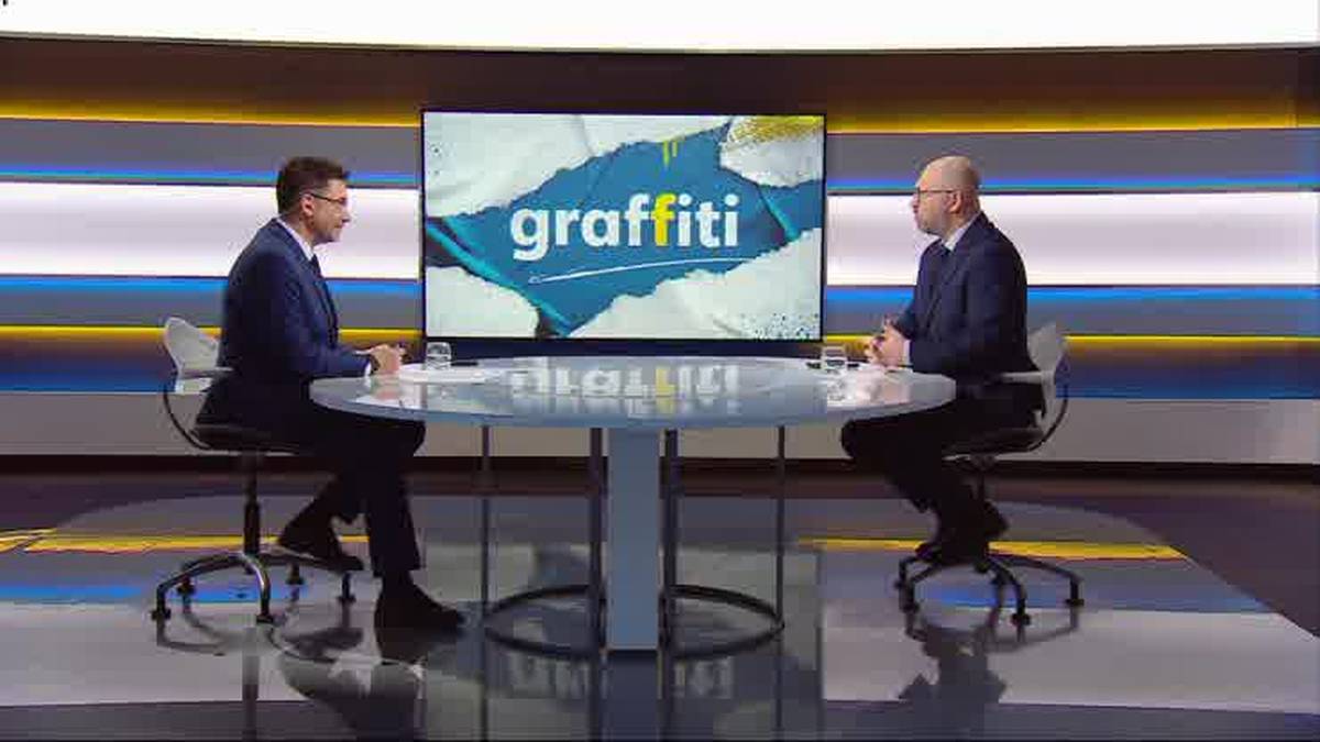 Dw&oacute;ch mężczyzn siedzi przy okrągłym stole w studiu telewizyjnym, naprzeciwko nich znajduje się duży ekran z napisem "graffiti".