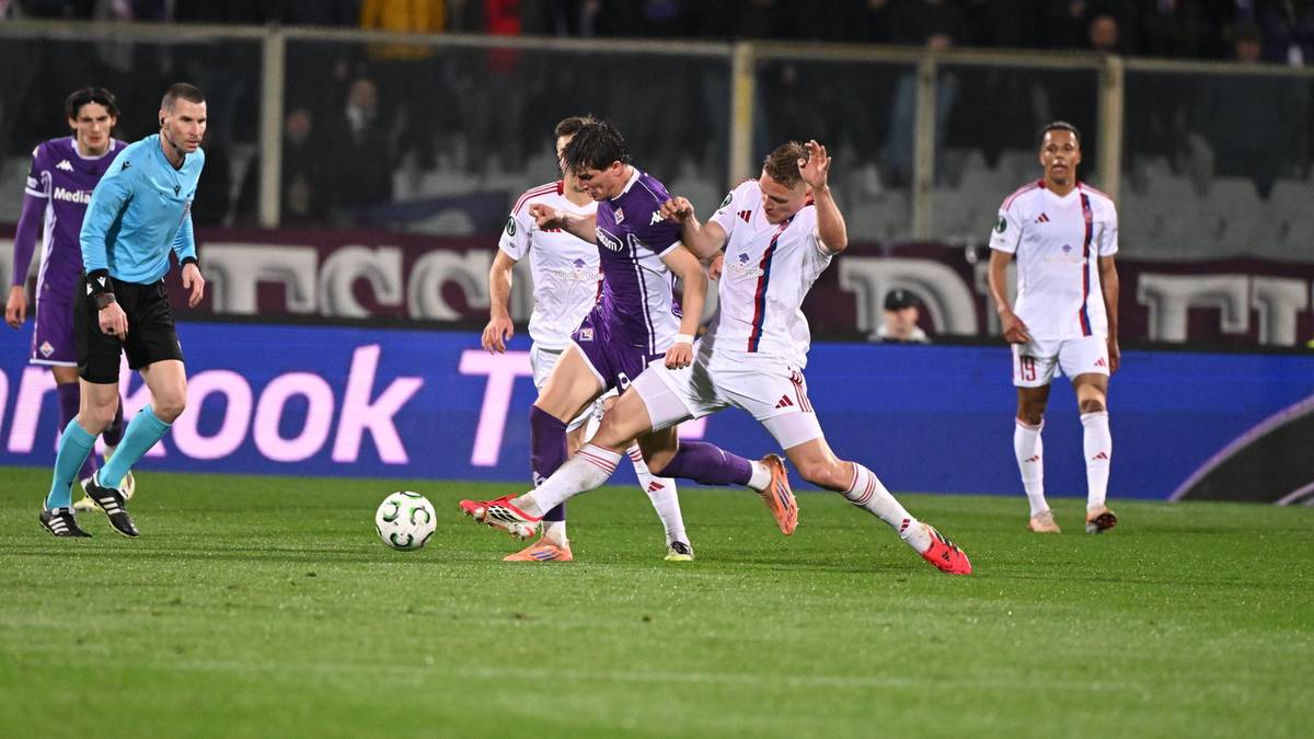 Piłkarze w białych i fioletowych strojach walczą o piłkę na boisku piłkarskim, z publicznością na trybunach w tle.