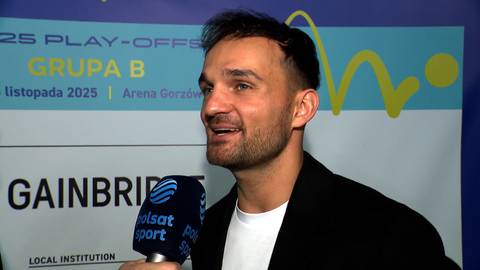 Portret Bartosza Zmarzlika podczas wywiadu. Uśmiechnięty mężczyzna z krótkimi włosami i zarostem, ubrany w białą koszulkę i czarną marynarkę, trzyma mikrofon Polsat Sport.