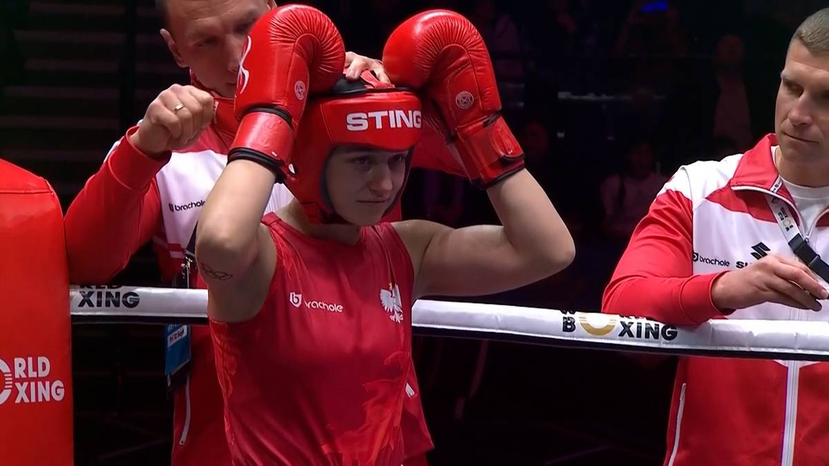 Bokserka w czerwonym stroju i rękawicach bokserskich poprawia hełm ochronny. Za nią stoją trenerzy.