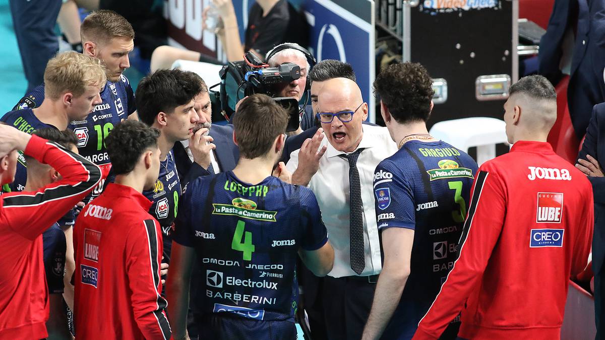 Siatkarze w czerwonych i granatowych strojach sportowych otaczają trenera podczas przerwy w meczu.