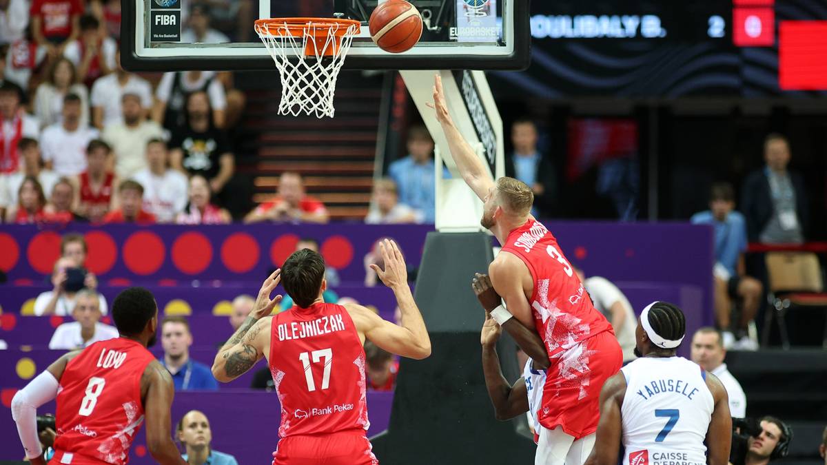 Gracz w czerwonej koszulce z numerem 17 rzuca piłkę do kosza, a gracz w czerwonej koszulce z numerem 8 próbuje go zablokować.