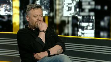 Mężczyzna z zarostem i tatuażem na przedramieniu siedzi w studiu telewizyjnym, zamyślony.