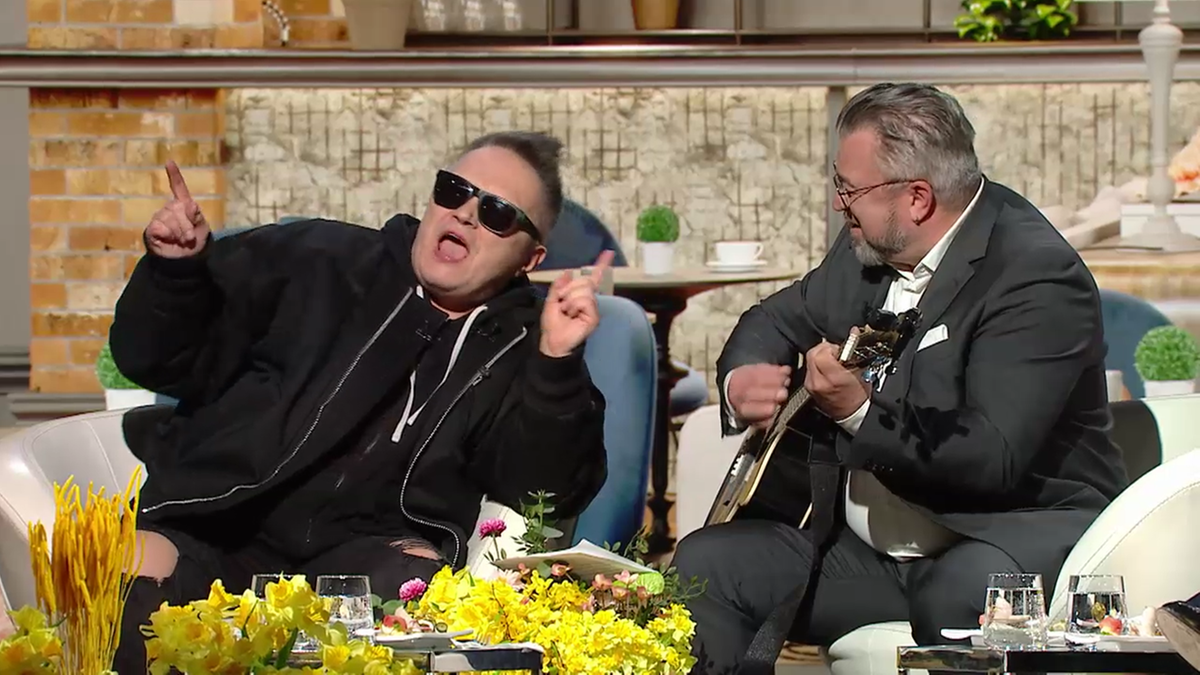 Muniek Staszczyk w okularach przeciwsłonecznych i czarnej bluzie śpiewa z uniesionymi rękami, podczas gdy Roman Kosecki siedzi obok i gra na gitarze.