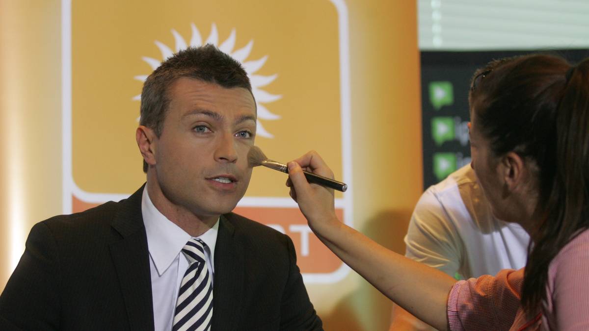 Mężczyzna w czarnym garniturze i białej koszuli z krawatem dostaje makijaż od kobiety z pędzlem do makijażu.