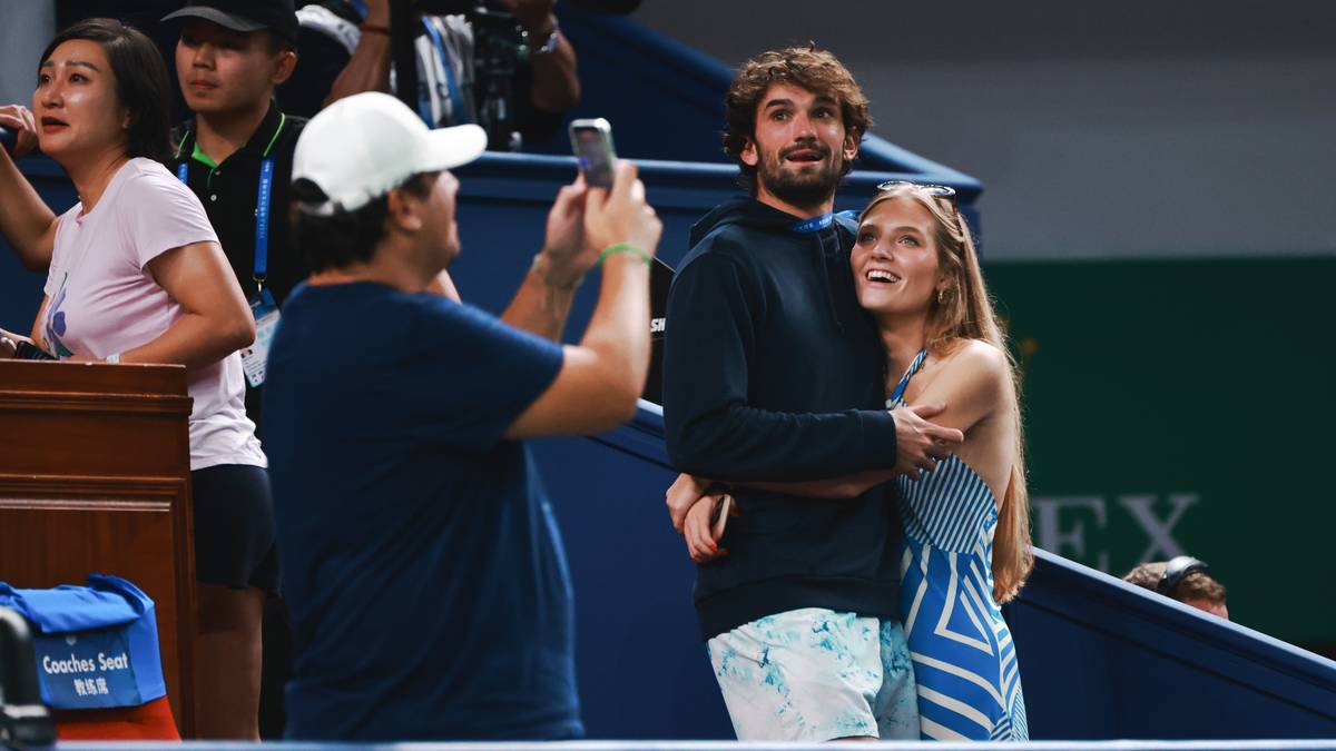 Para przytulająca się na trybunach podczas meczu tenisowego. Mężczyzna z zarostem i brązowymi włosami obejmuje kobietę. Kobieta w opasce na włosach odwzajemnia uścisk.