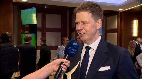 Łukasz Sekuła w garniturze odpowiada na pytanie dziennikarza trzymającego mikrofon z logo Polsat Sport. W tle widać inne osoby oraz ekrany z zieloną treścią.