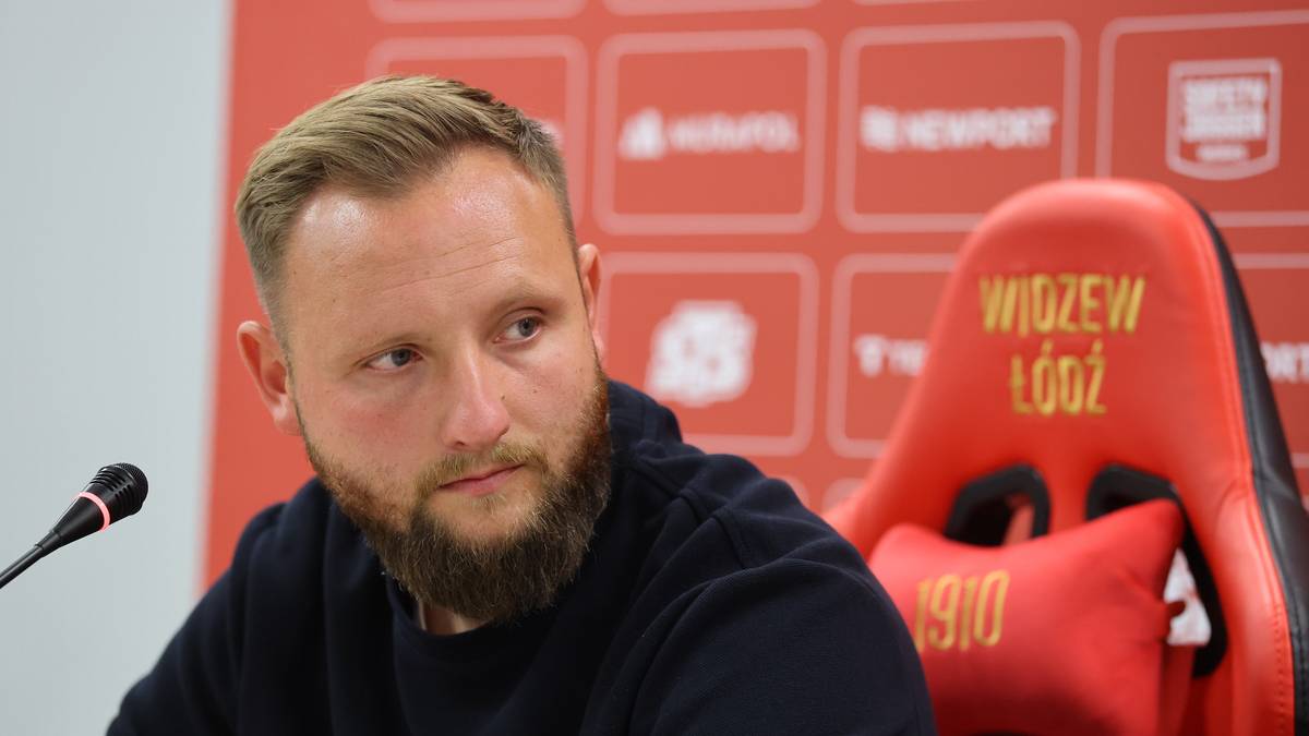 Mężczyzna z brodą, ubrany w ciemny sweter, siedzi przy mikrofonie. Za nim widać czerwony fotel z napisem "WIDZEW Ł&Oacute;DŹ" i datą "1910".