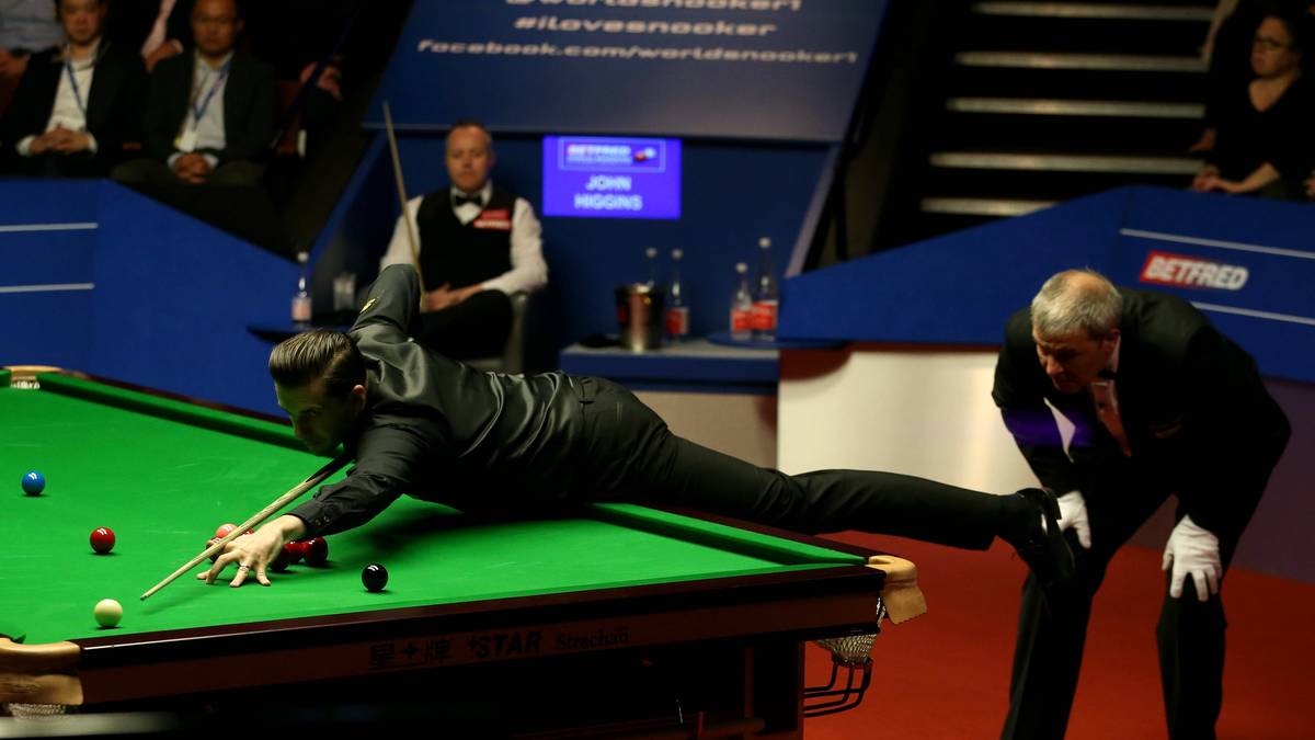 Gracz snookera w czarnym stroju pochyla się nad stołem i wykonuje uderzenie. Obok niego stoi sędzia w białej koszuli i czarnym krawacie.
