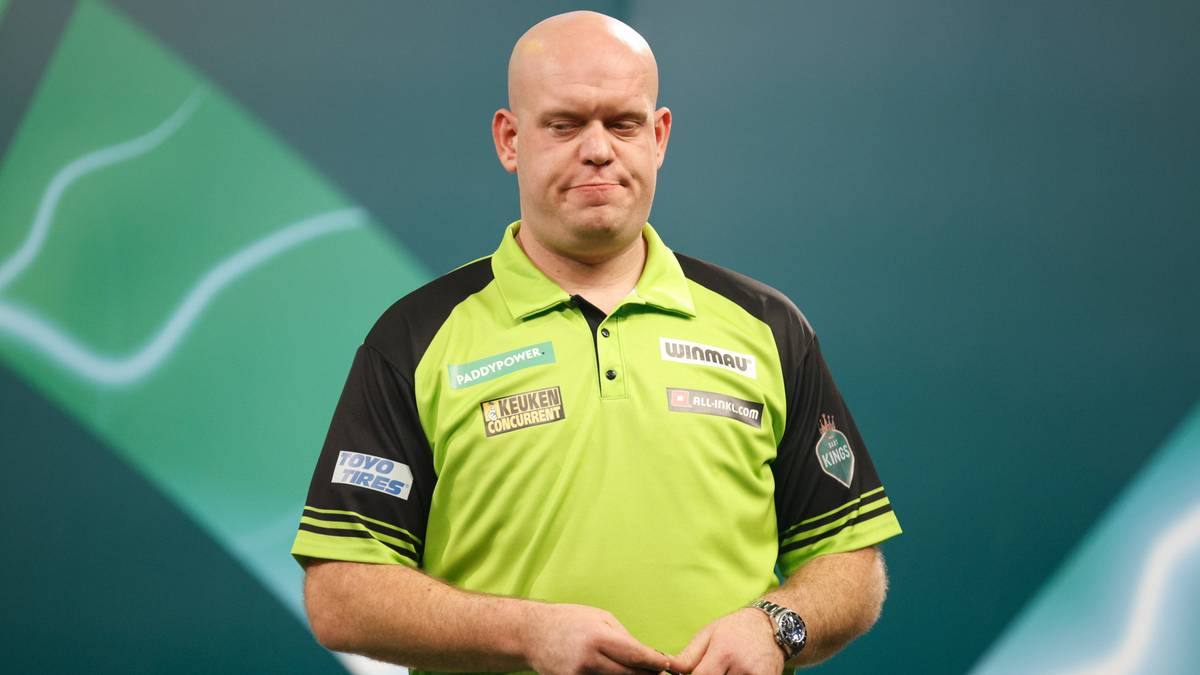 Mężczyzna w zielono-czarnej koszulce polo z logo sponsora, ubrany w str&oacute;j sportowy do gry w darty, stoi przed zielonym tłem z geometrycznymi wzorami.