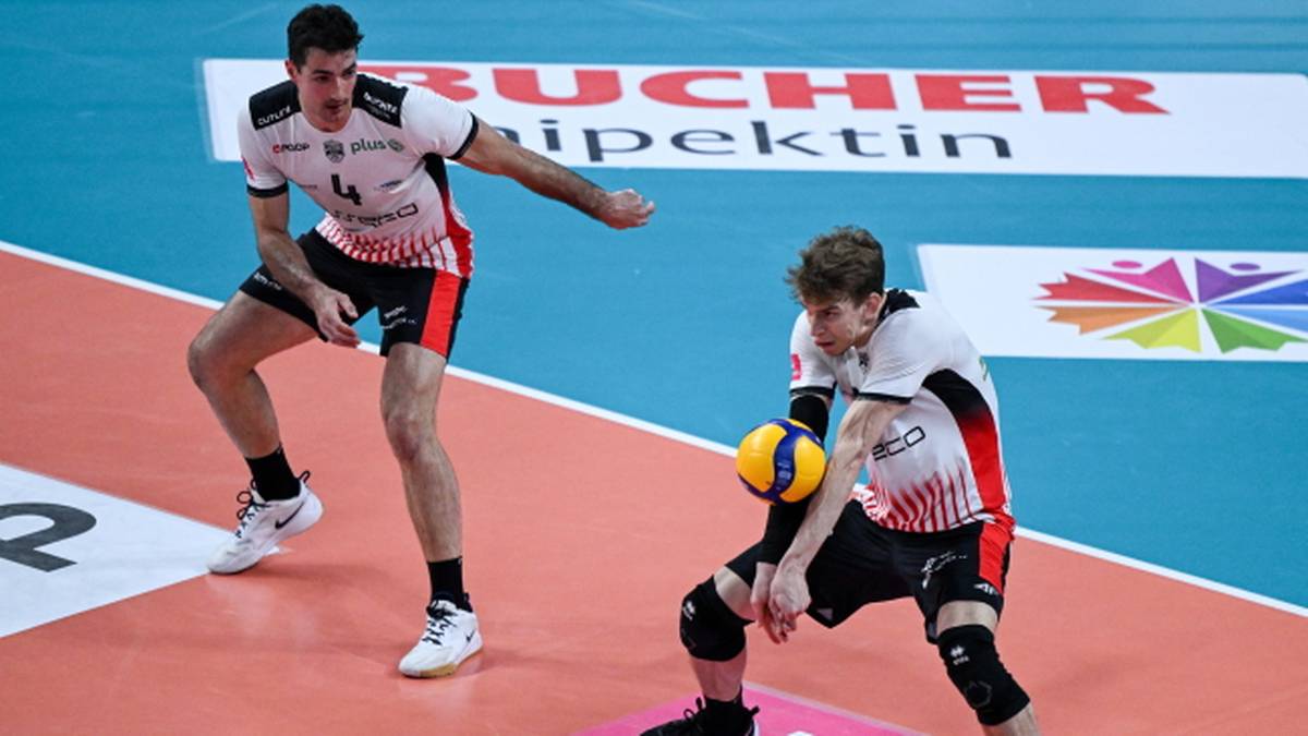 Siatkarz w biało-czerwonym stroju z numerem 14 na plecach wykonuje zagrywkę, a drugi siatkarz w podobnym stroju odbiera piłkę z&oacute;łto-niebieską piłkę.