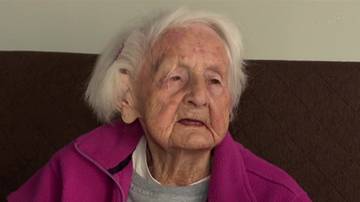 Portret starszej kobiety z siwymi włosami i pomarszczoną twarzą, ubraną w r&oacute;żową kurtkę.