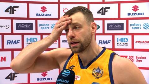 Mężczyzna w ż&oacute;łtej koszulce koszykarskiej trzyma się za czoło prawą ręką, wyrażając zmartwienie lub zaw&oacute;d. Za nim znajduje się ściana z logo sponsor&oacute;w.