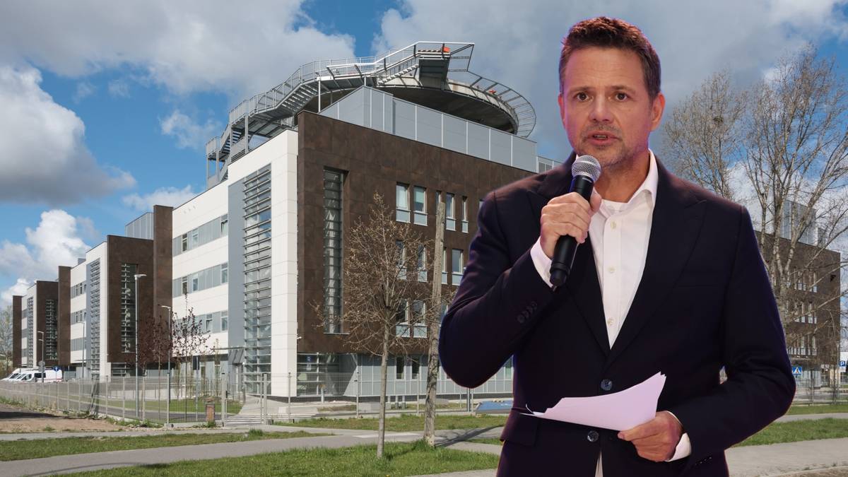 Mężczyzna w garniturze przemawia do mikrofonu na tle budynku szpitala.