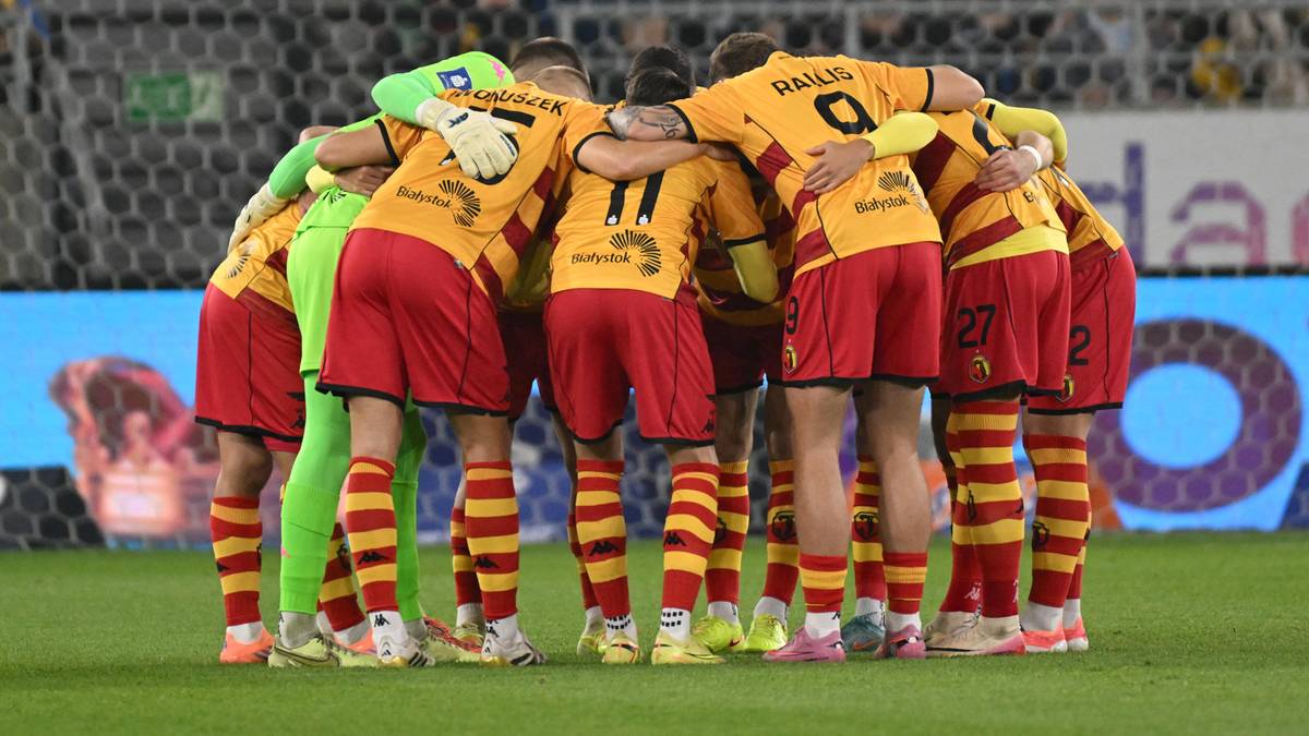 Piłkarze w żółto-czerwonych strojach stoją w kółku na boisku.