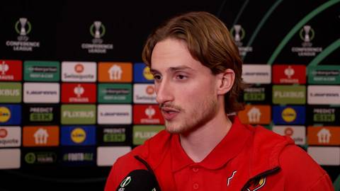 Mężczyzna z jasnymi włosami, ubrany w czerwoną koszulkę, udziela wywiadu na tle ściany z logo Ligi Konferencji Europy i sponsor&oacute;w.