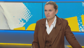 Kobieta w eleganckim brązowym garniturze siedzi na tle ekranu z kolorowymi abstrakcyjnymi wzorami.