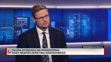 Mężczyzna w okularach i garniturze siedzi przed ekranem telewizyjnym.