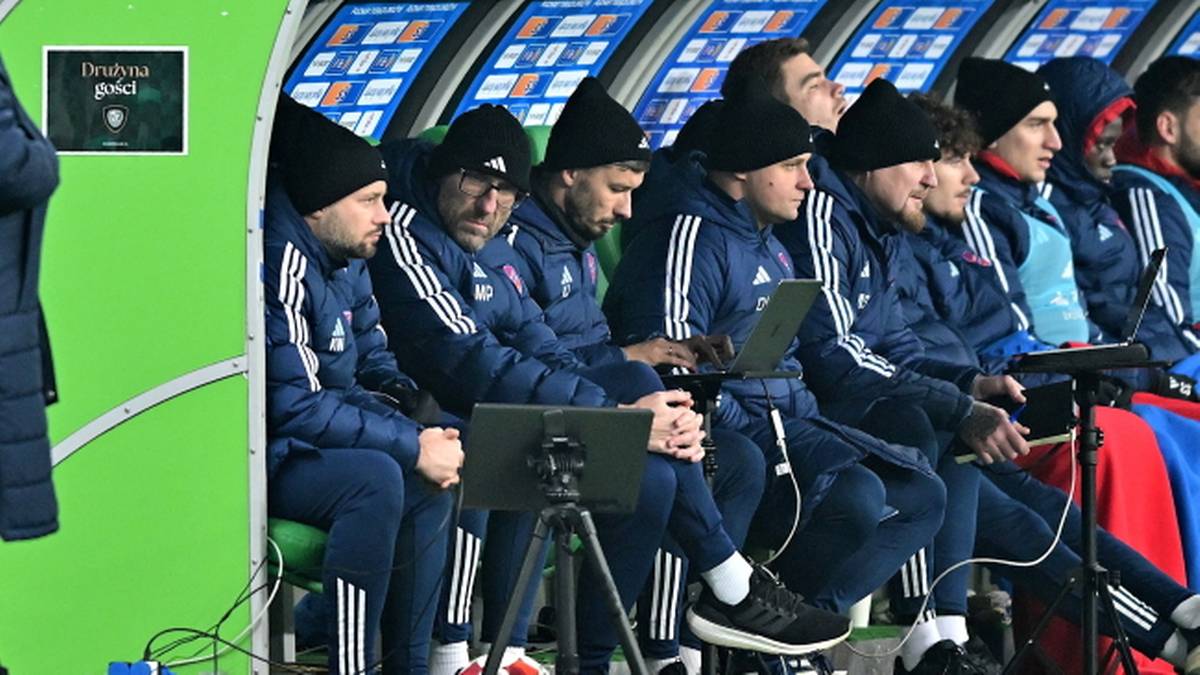 Trenerzy siedzący na ławce rezerwowych podczas meczu piłkarskiego, jeden z nich korzysta z laptopa.