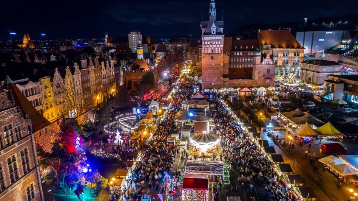 Nocny widok na zatłoczony jarmark bożonarodzeniowy w historycznym centrum miasta, z licznymi straganami, iluminacjami i budynkami.