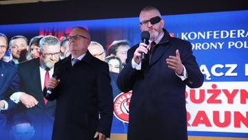 Dwóch mężczyzn w płaszczach stojących przed ekranem z napisem "Konfederacja Korony Polskiej"