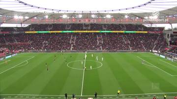 Widok z góry na boisko piłkarskie podczas meczu Bayer Leverkusen - Bayern Monachium. Na zielonej murawie, podzielonej białymi liniami, znajduje się dwóch zawodników w białych strojach i kilku w czerwonych. Trybuny wokół boiska są wypełnione kibicami w ciemnych ubraniach.