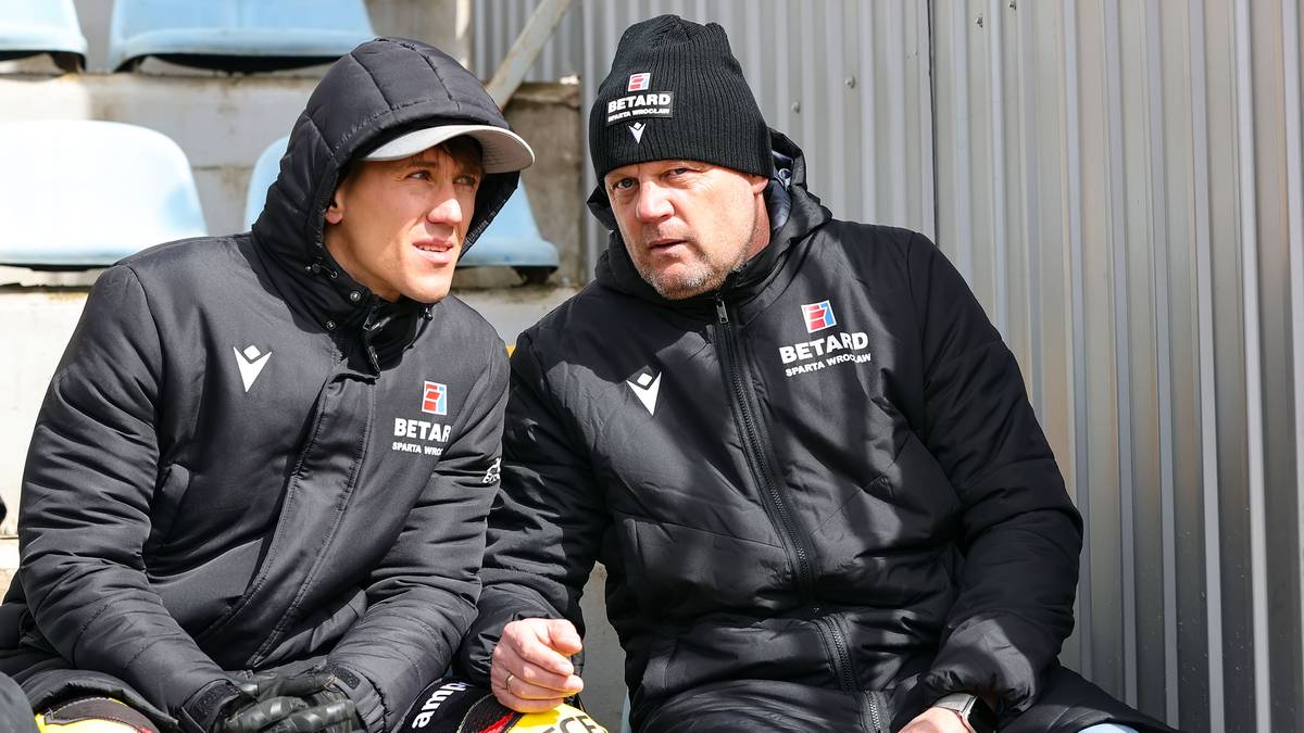 Dwaj mężczyźni w czarnych kurtkach i czapkach, siedzą obok siebie. Jeden z nich, młodszy, w kapturze, patrzy w bok z zaniepokojoną miną. Starszy mężczyzna, z krótkimi siwymi włosami, ubrany w czapkę z logo, mówi coś do młodszego, z wyraźnym wzburzeniem na twarzy. Obydwaj mają na sobie kurtki z widocznym logo "BETARD"