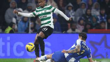 Piłkarz w zielono-białej koszulce pr&oacute;buje odebrać piłkę leżącemu na trawie zawodnikowi w niebieskiej koszulce.