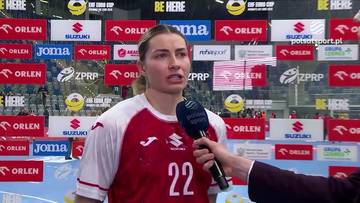 Młoda kobieta w białej koszulce sportowej z czerwonymi rękawami i numerem 22 na plecach, trzyma mikrofon i odpowiada na pytania dziennikarza. W tle znajdują się ściany z logotypami sponsor&oacute;w.