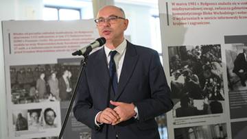 Mężczyzna w garniturze i okularach przemawia przy mikrofonie, obok niego wystawa z dokumentami i zdjęciami.
