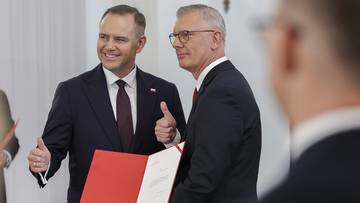 Dwóch mężczyzn w garniturach, jeden z nich uśmiecha się i pokazuje kciuk w górę, trzymając czerwony folder.