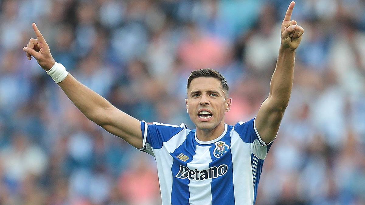 Piłkarz w niebiesko-białej koszulce z białymi pasami, z podniesionymi palcami wskazującymi w górę, na tle trybun wypełnionych kibicami.