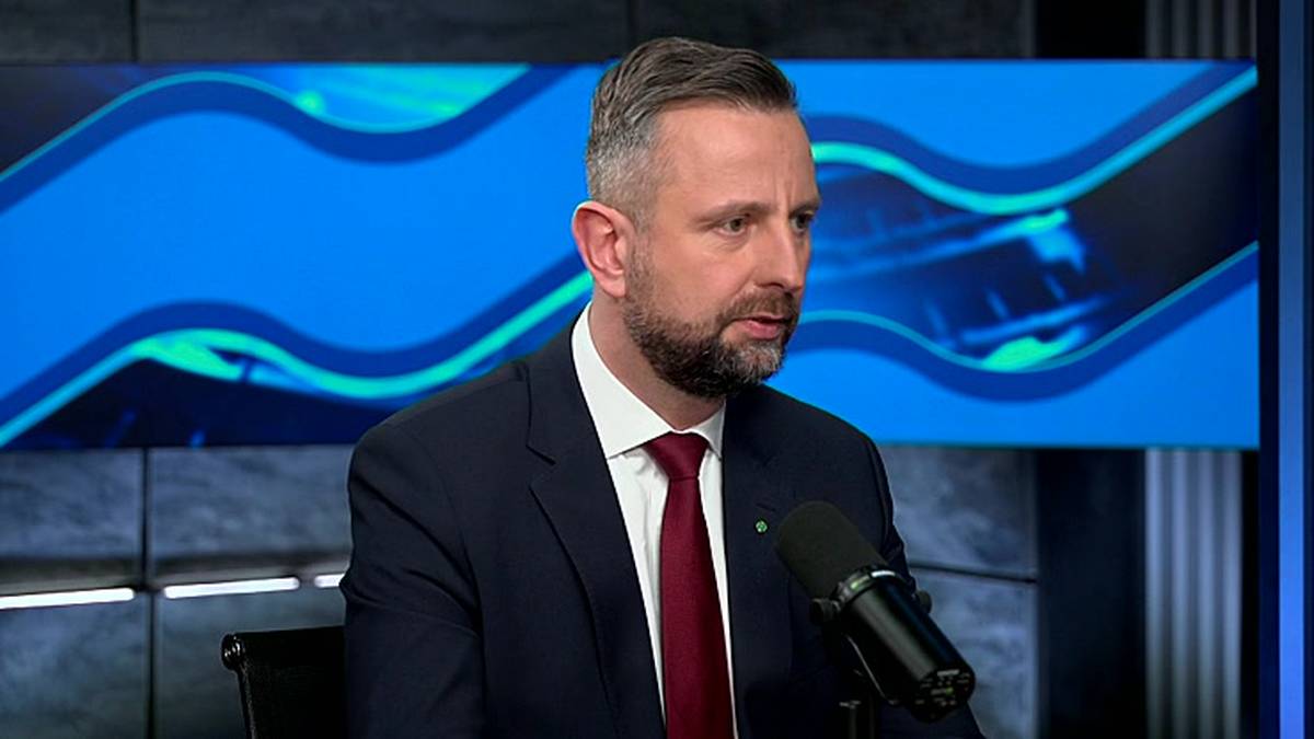 Mężczyzna w garniturze i krawacie siedzi przed mikrofonem, na tle niebieskiej grafiki z falami.