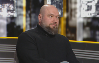 Mężczyzna z brodą w czarnym golfie siedzi przed kamerą.